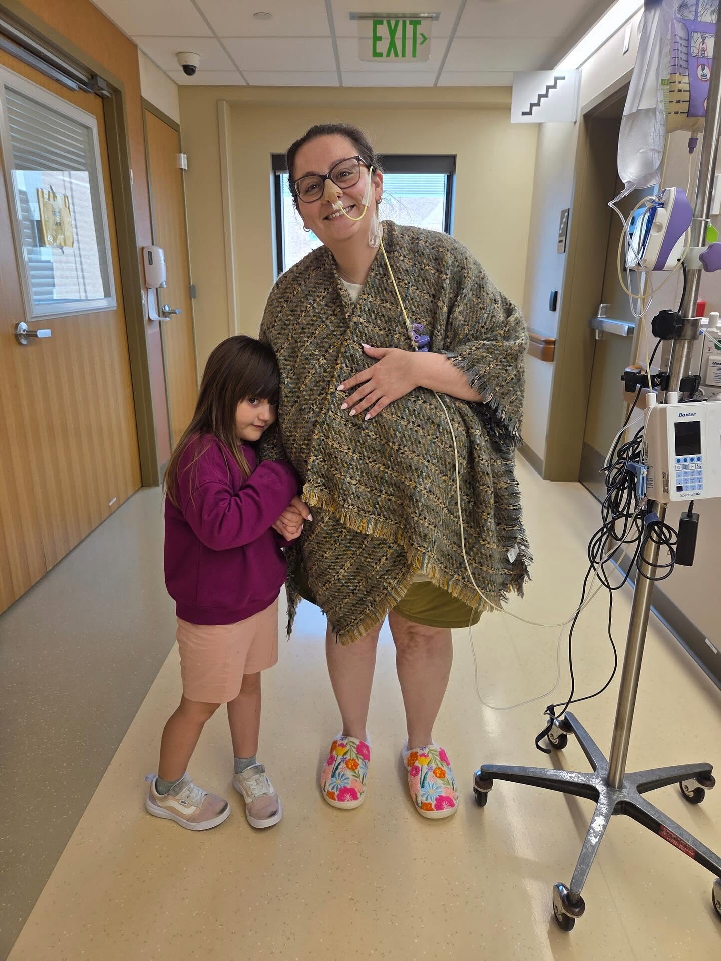 Woman and girl standing in a hospital corridor, woman wearing a patterned shawl and colorful shoes, girl in pink hoodie and shorts.
