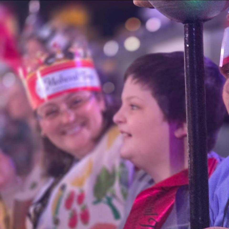A smiling woman and a boy are at a festive event, with colorful decorations and blurred lights in the background.