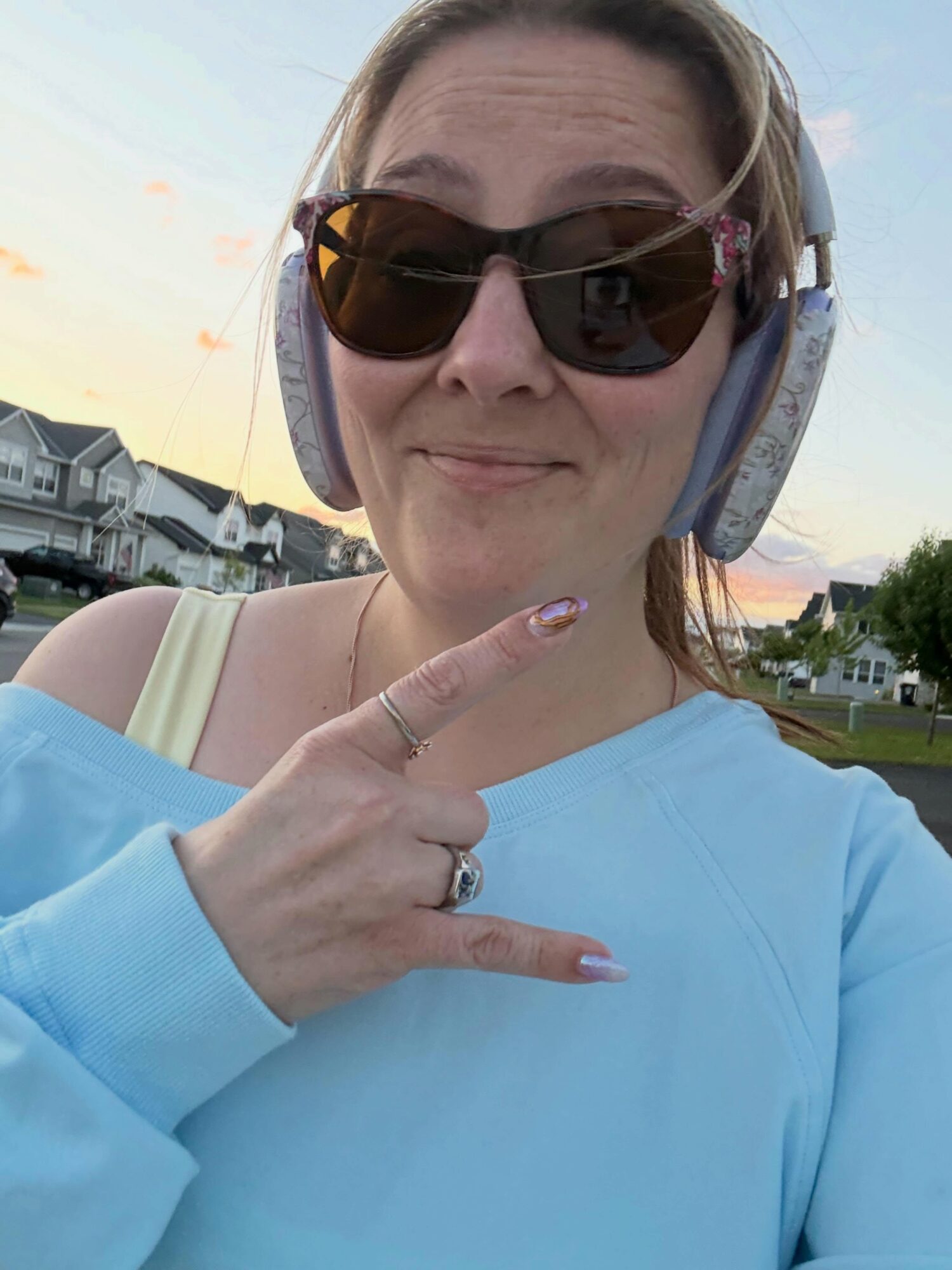 Woman wearing sunglasses and headphones, pointing to her chest with her right hand, outdoors during sunset.