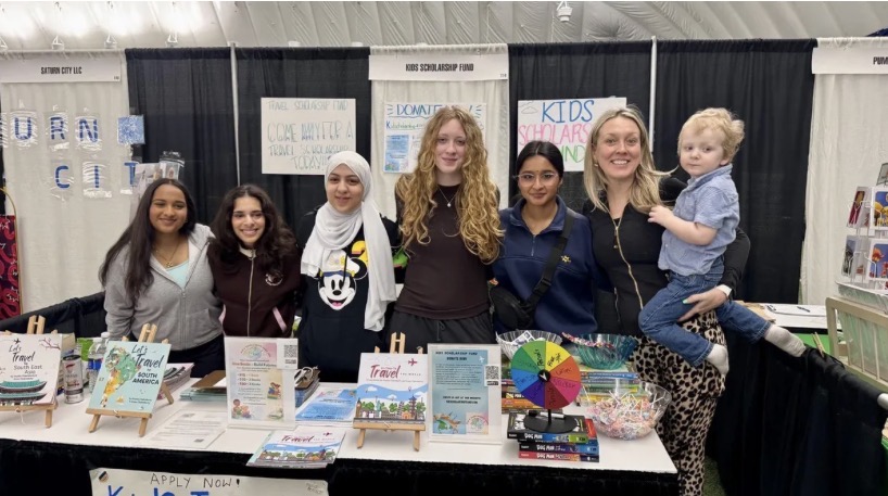Group of seven people, including children and adults, standing behind a table with books and colorful materials at an indoor event.