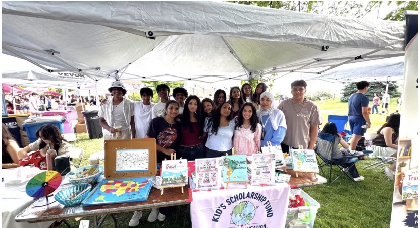 Group of people standing behind a table with colorful displays and signs at an outdoor event under a tent.