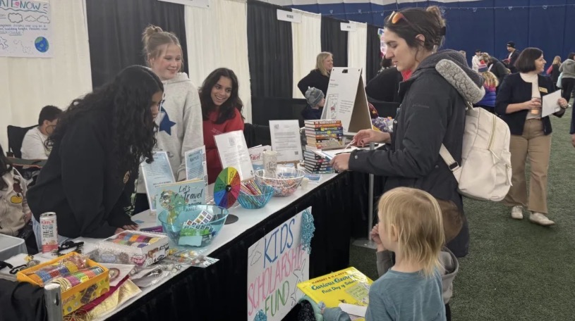Children and adults at a booth with colorful items and signs, indoors at an event or fair.