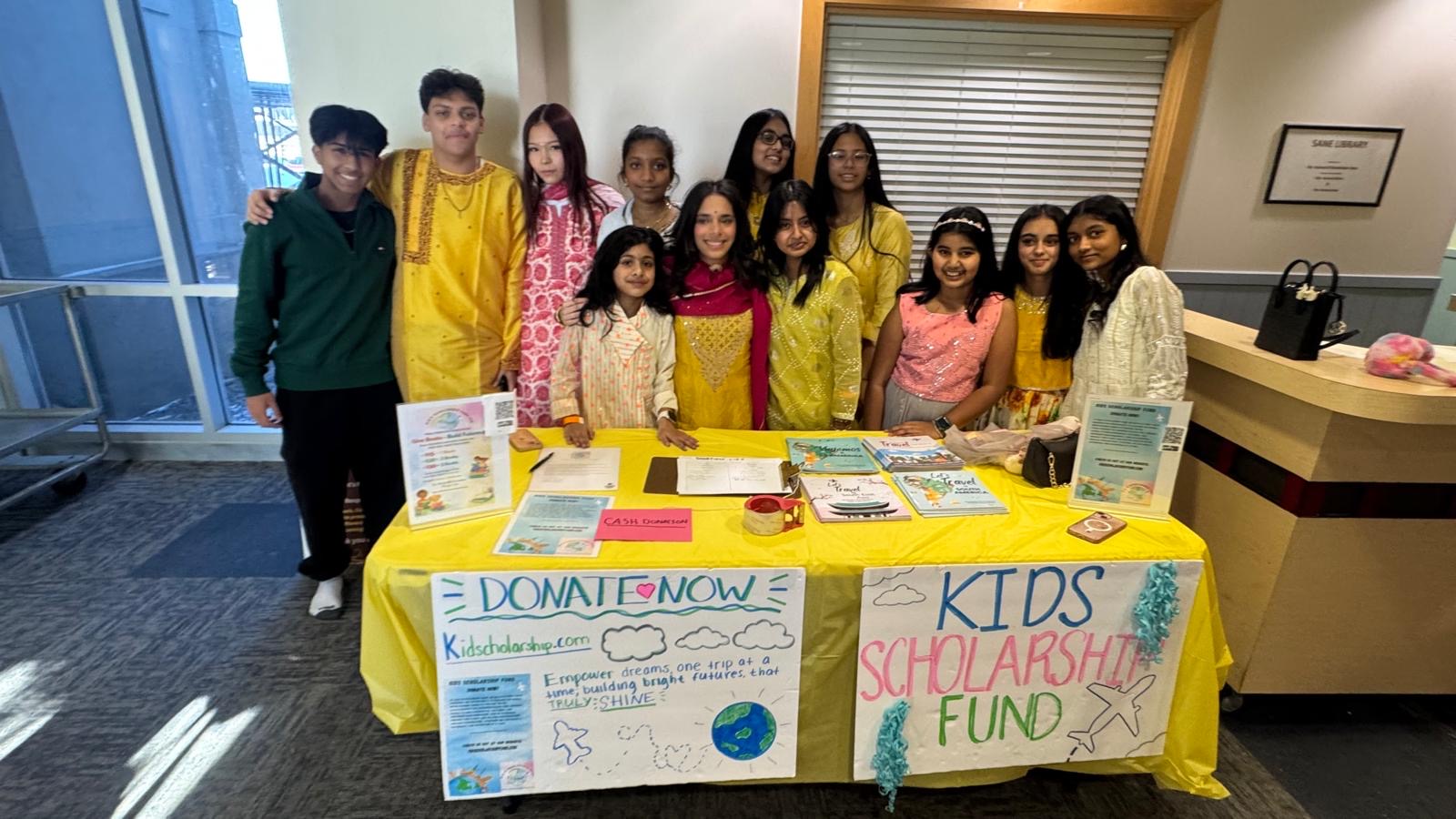 Group of children standing behind a colorful fundraising table with posters, in an indoor setting.