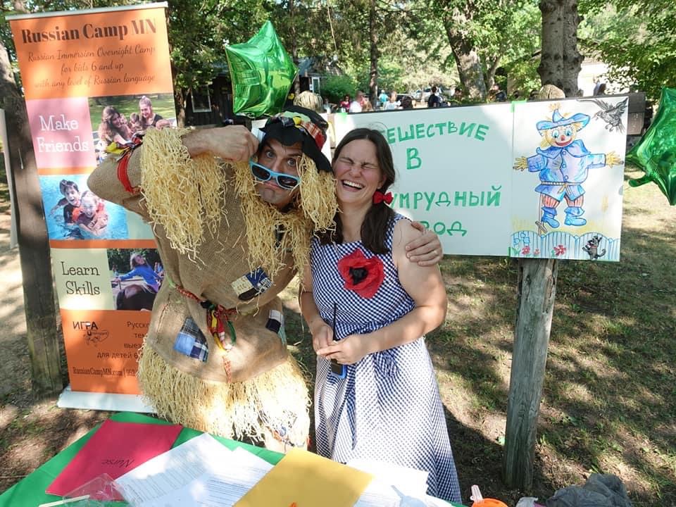 Two women smiling outdoors near a colorful sign and poster, with trees and grass in the background.