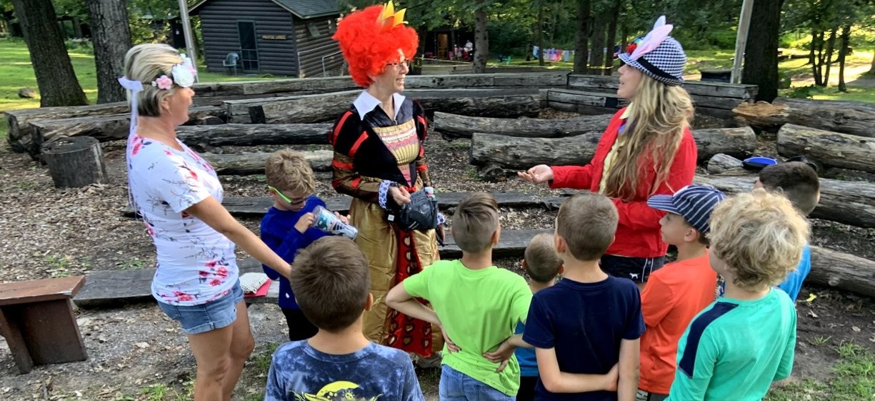 Group of children and two adults outdoors, with one adult dressed as a clown and another as a witch, engaging children.