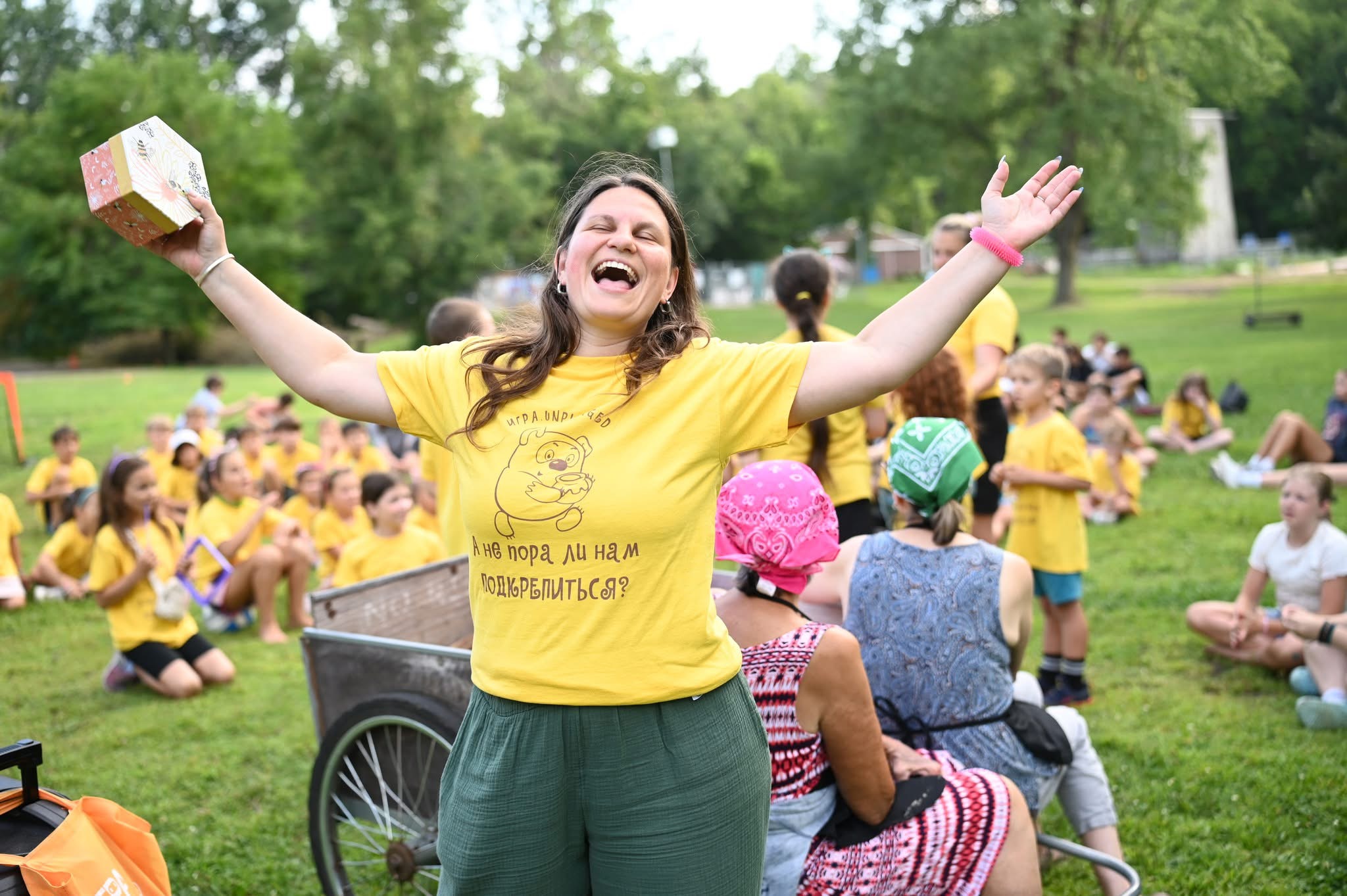 Woman smiling with arms raised outdoors, surrounded by children in yellow shirts, some sitting on grass, trees in background.