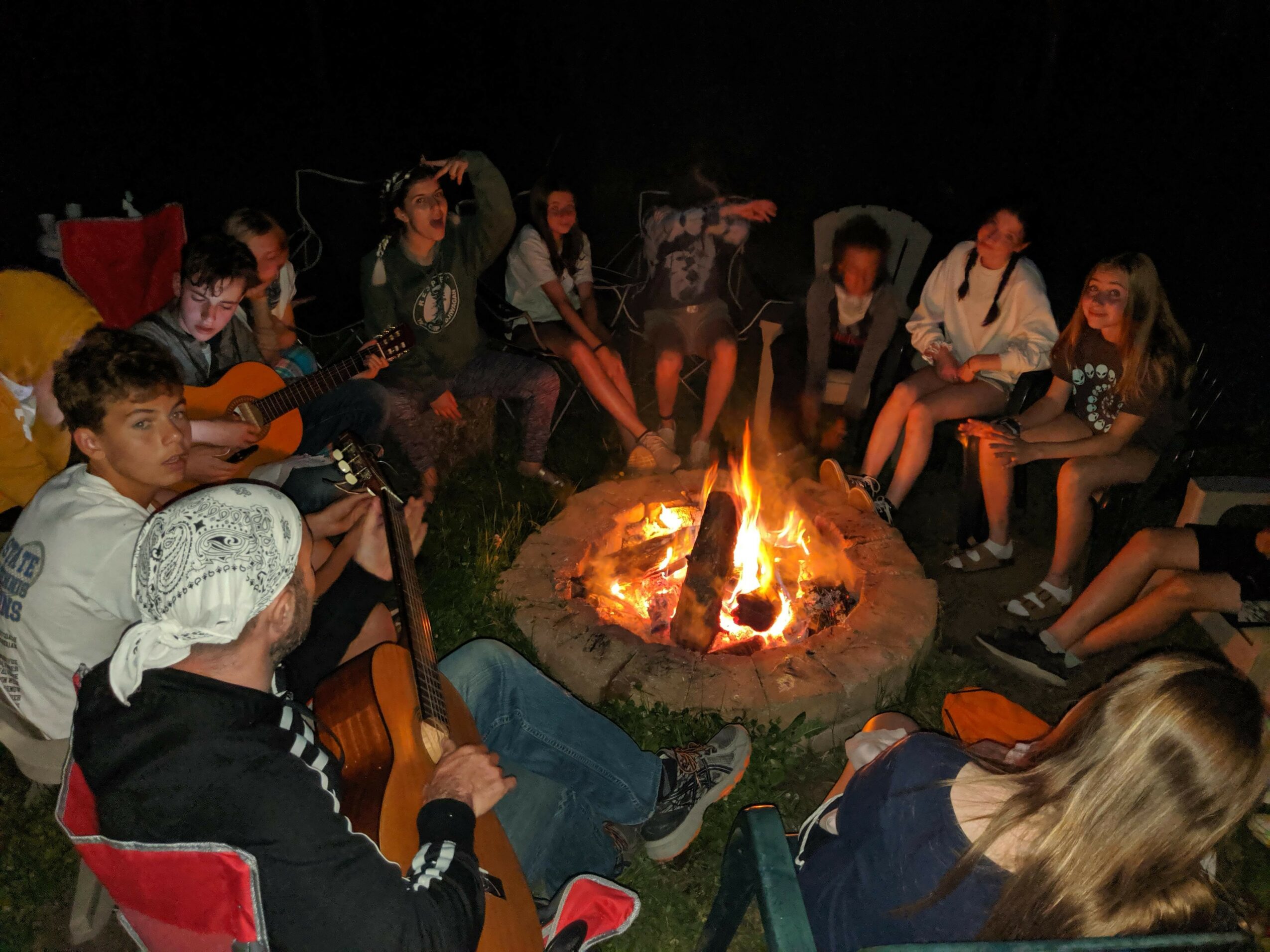Group of people sitting around a campfire at night, some playing guitars, others talking and relaxing.