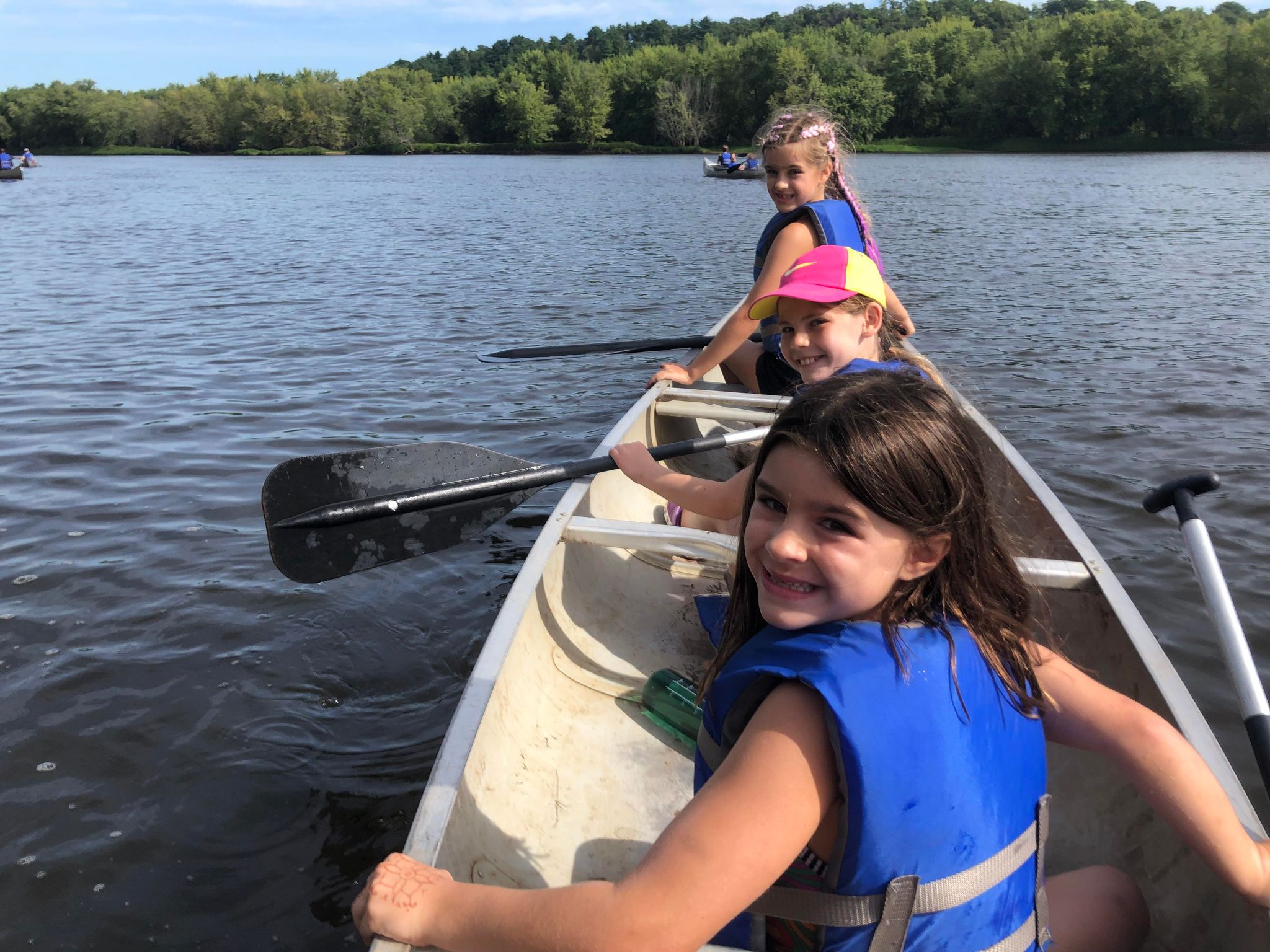 Three children in a boat on a lake, smiling and paddling, with trees in the background.