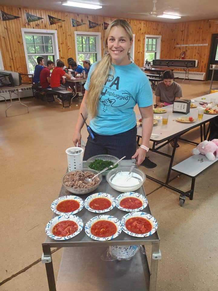 Young woman with long blonde hair smiling behind a table with seven plates of food, in a wooden room with windows and people in the background.