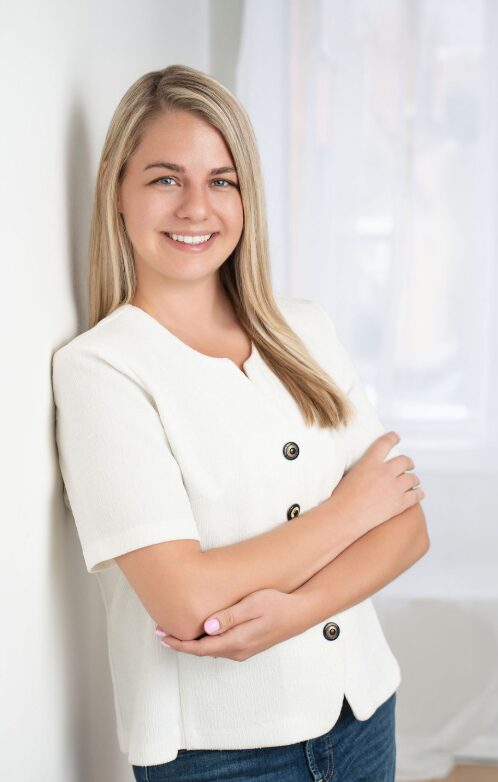 Young woman with blonde hair smiling, wearing a white top, standing indoors near a window.