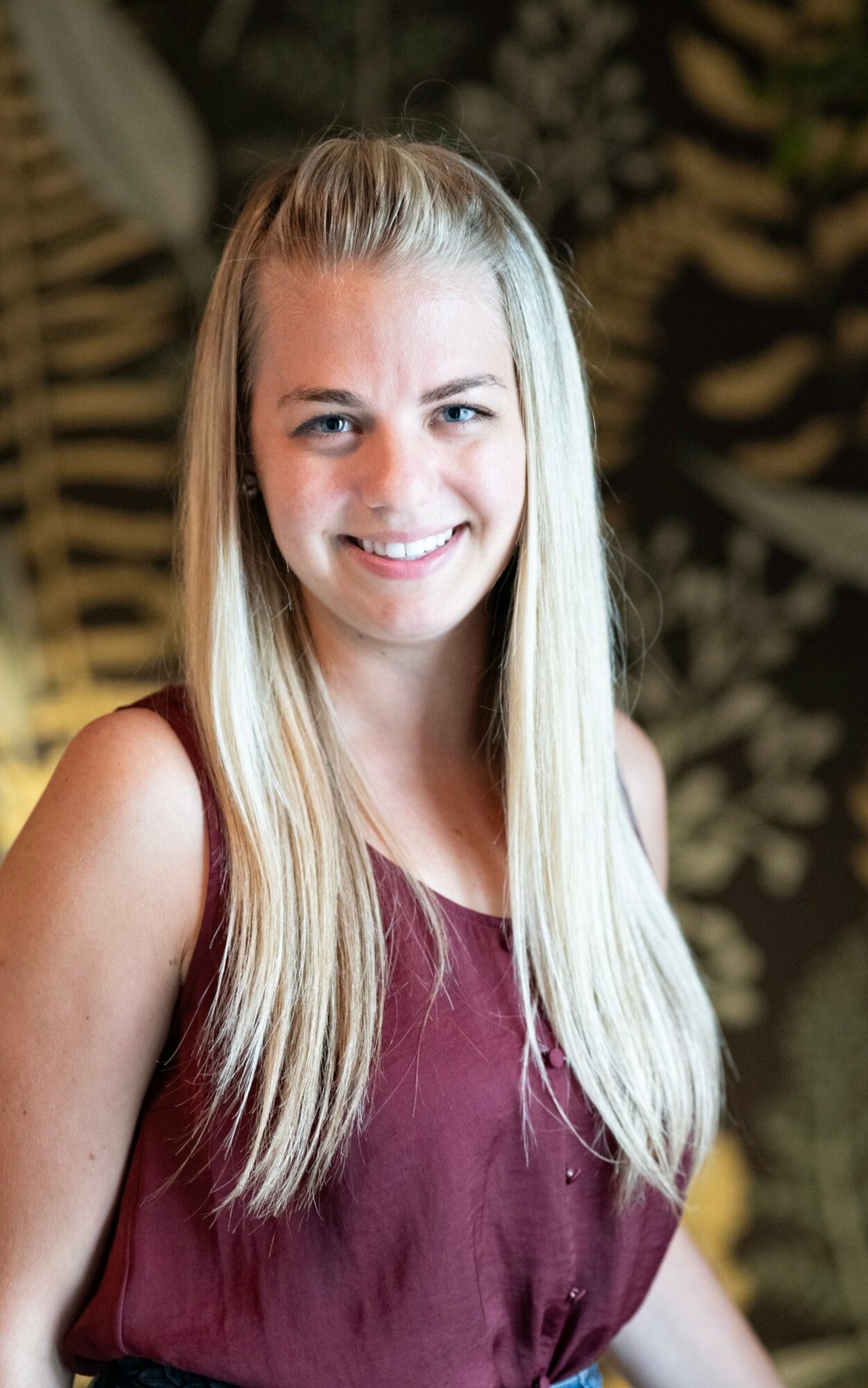 Young woman with long blonde hair smiling, wearing a sleeveless maroon top, standing outdoors with blurred foliage background.