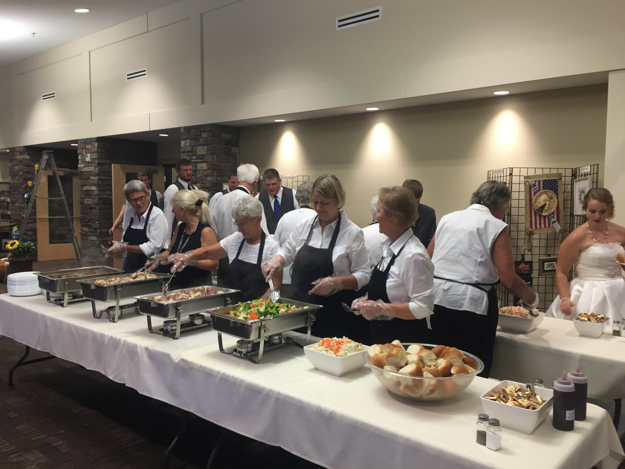 People in white shirts and black aprons serve food at a buffet table in a well-lit room.