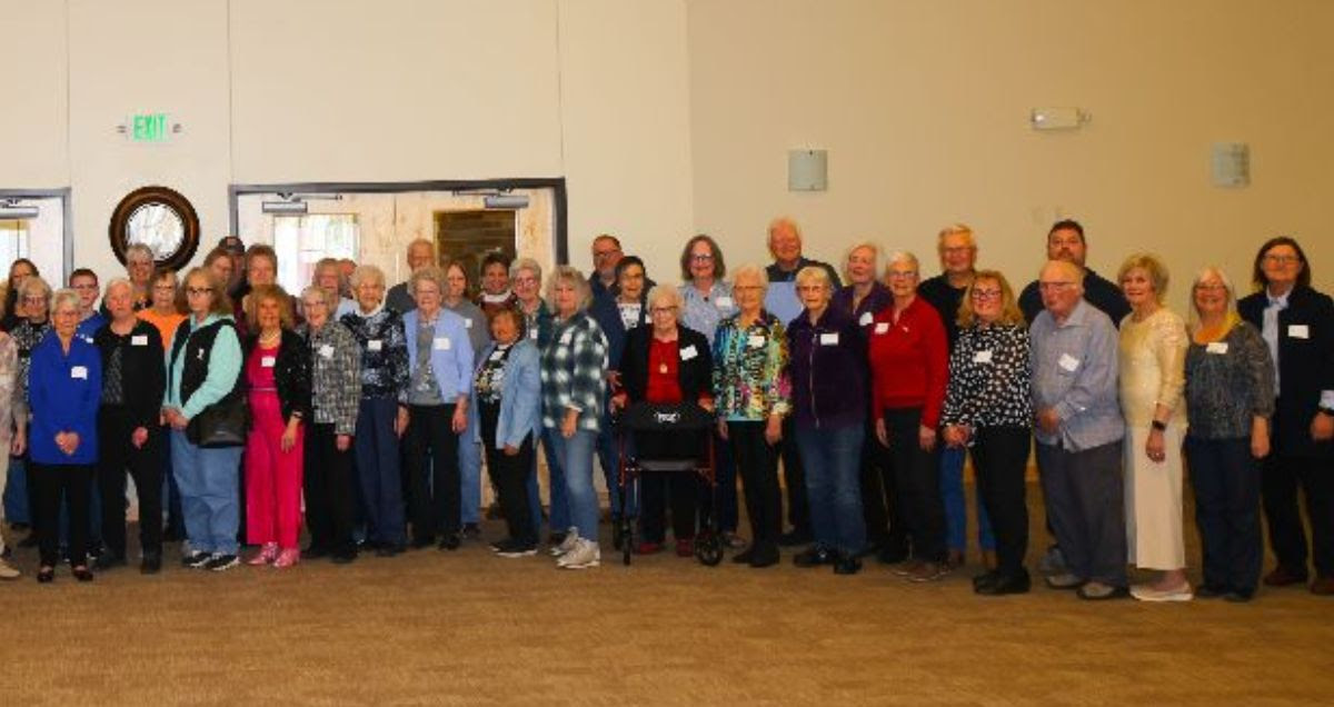 Group of people standing indoors in a large room, posing for a photo, with a beige wall and door behind them.