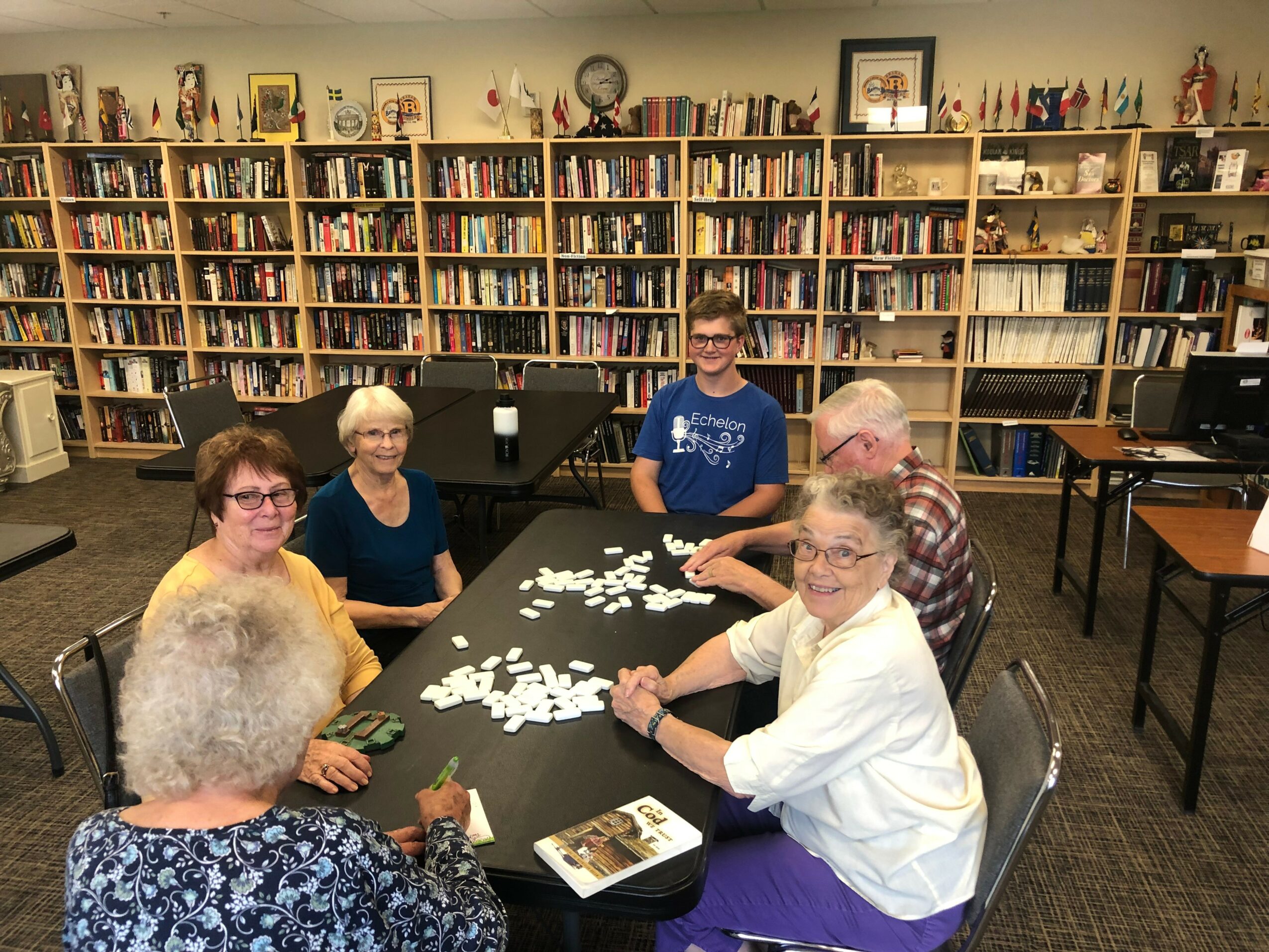 Six people sit around a table in a library, smiling and engaging in a game or activity, with bookshelves in the background.