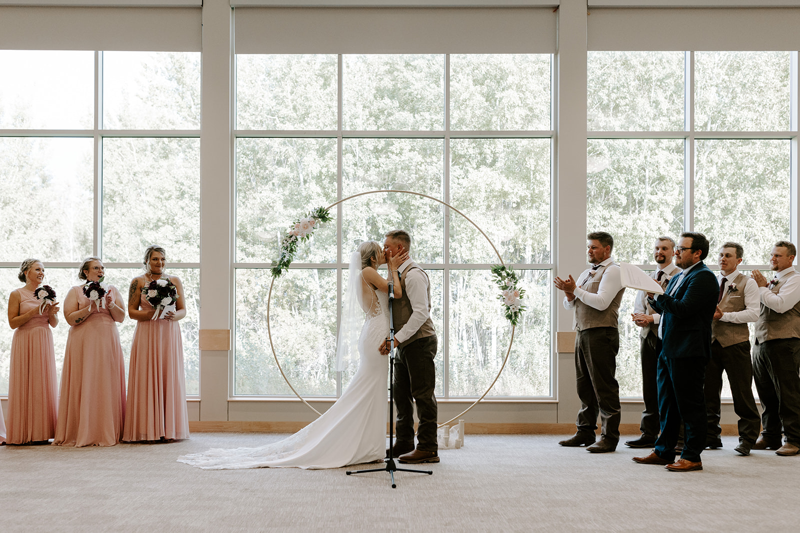 Bride and groom kiss in front of large windows, surrounded by wedding party and guests, during ceremony indoors.