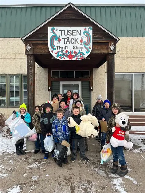 Group of children and adults standing outside a building with a sign that reads 'Tusen TACK Thrift Shop,' holding bags and stuffed animals.