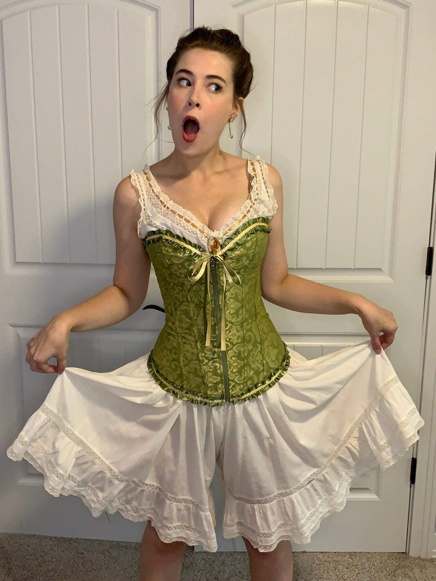 Young woman with surprised expression, wearing a green corset and white skirt, standing in front of white doors.