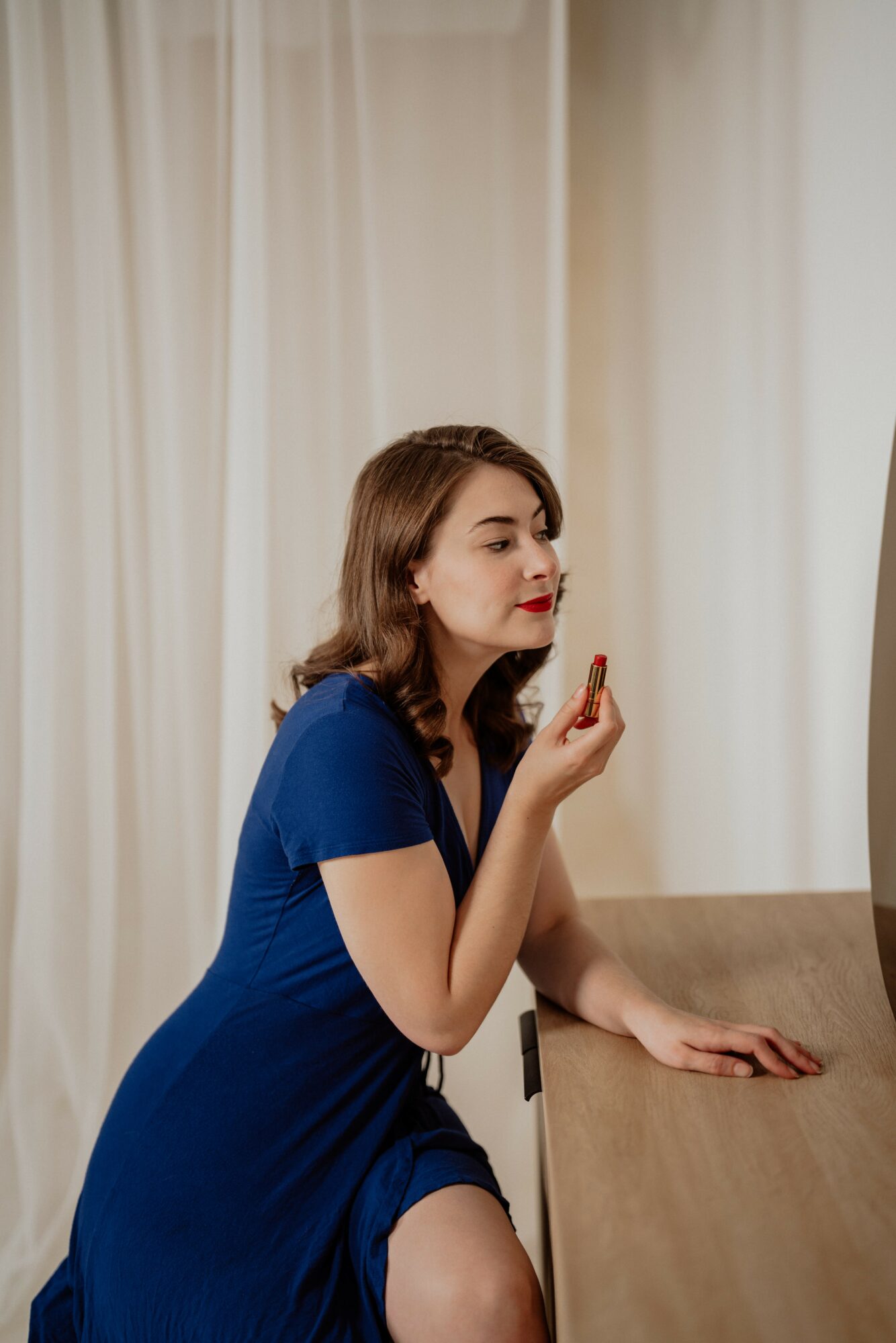 Woman in blue dress sitting at a desk, holding a lipstick near her face, with curtains in the background.