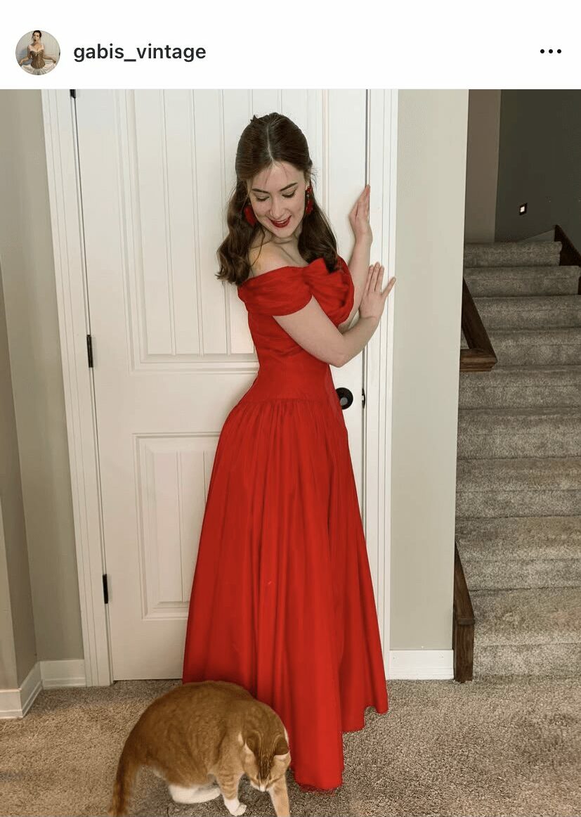 Young woman in red dress standing indoors with a small dog at her feet, door and staircase in background.