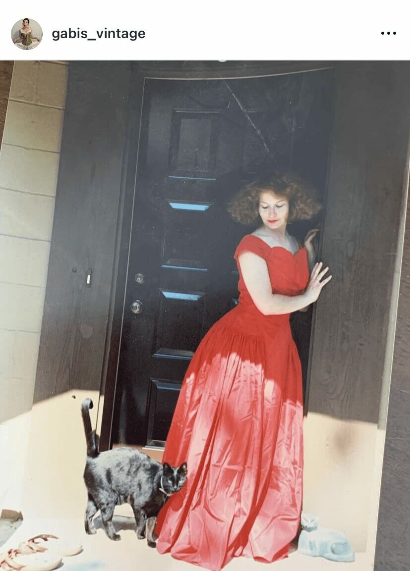 Woman in a red dress standing next to a small dog in front of a black door, with a white object nearby.