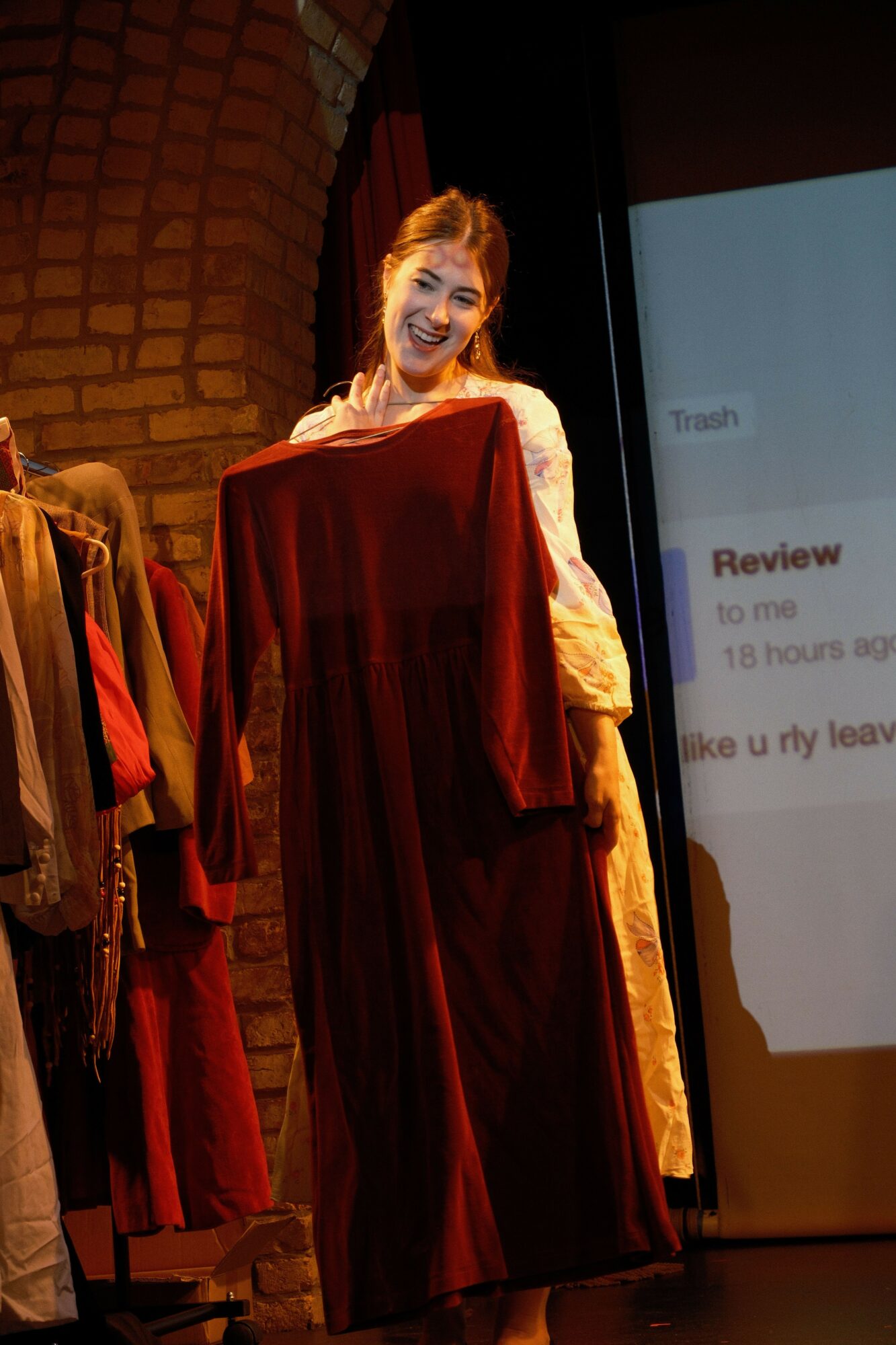 Person in a long red dress standing on stage with a brick wall background, speaking or performing.
