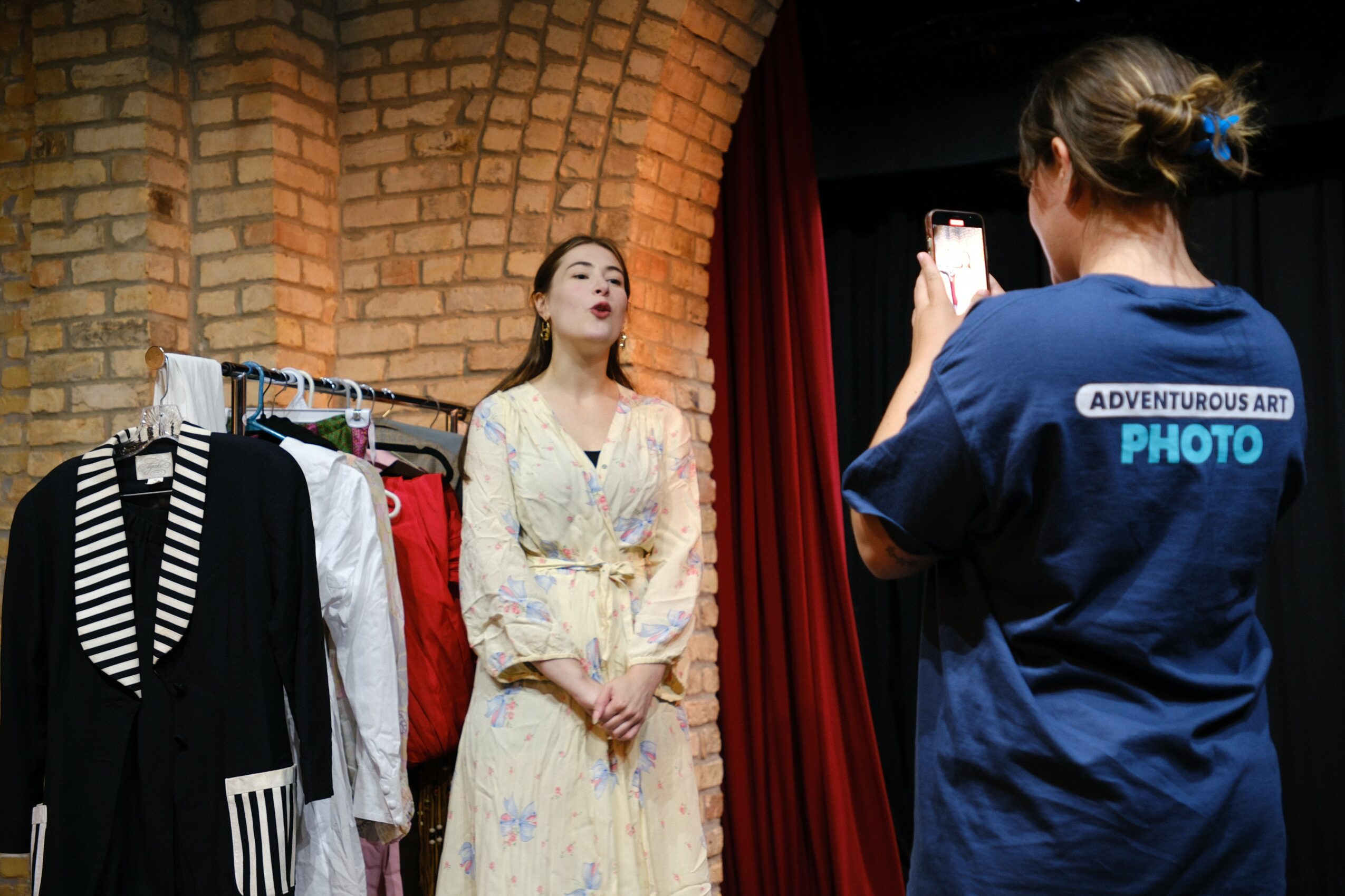 A woman in a floral dress stands against a brick wall while a person in a blue shirt takes a photo.