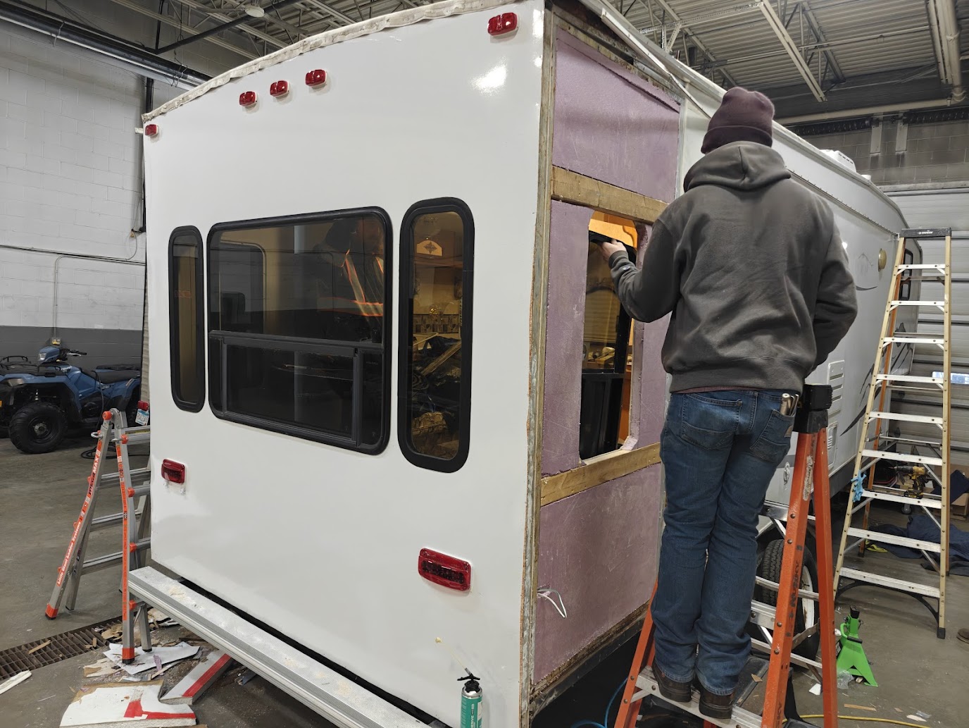 Person in gray hoodie and jeans working on a small white and purple trailer inside a warehouse.