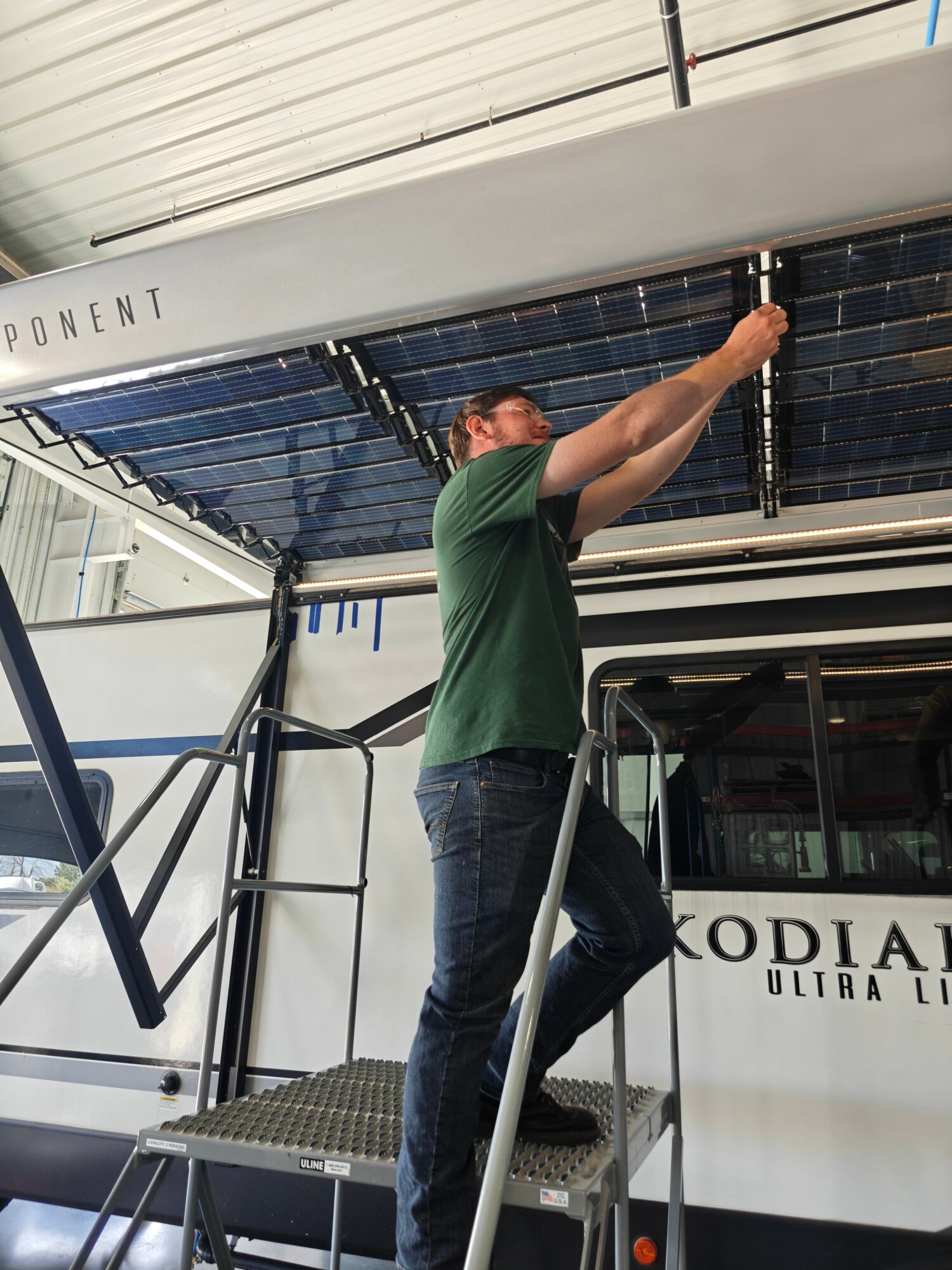 Person in green shirt and jeans working on boat, standing on a platform, reaching up to the boat's underside.