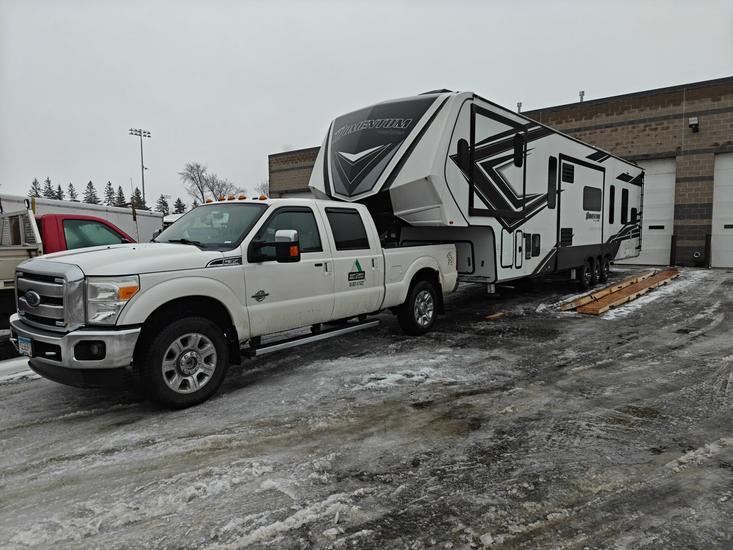 White pickup truck towing a large modern RV parked outside on a wet, snowy ground with a brick building in the background.