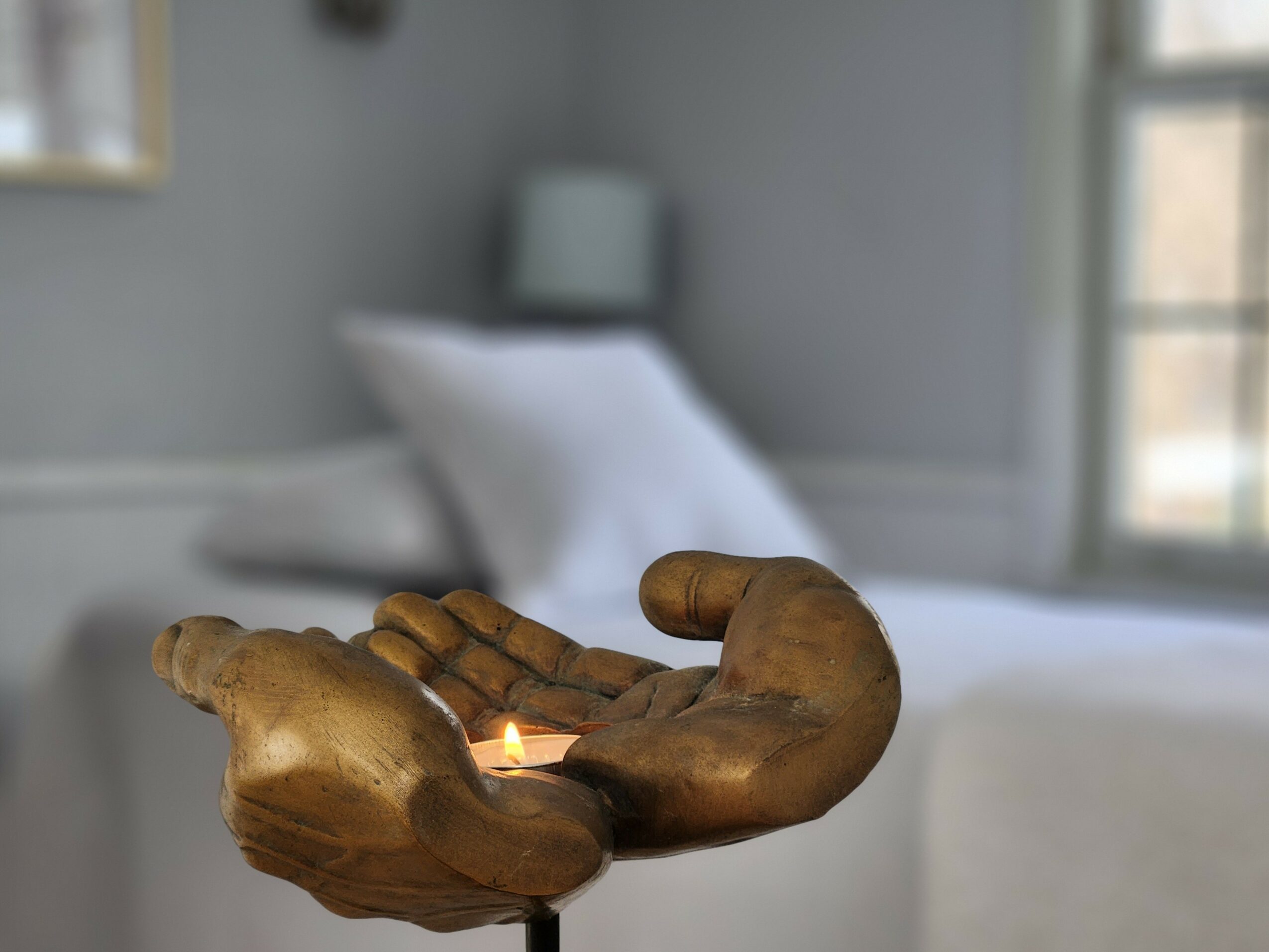 Two hands holding a small lit candle in a room with a bed and window in the background.