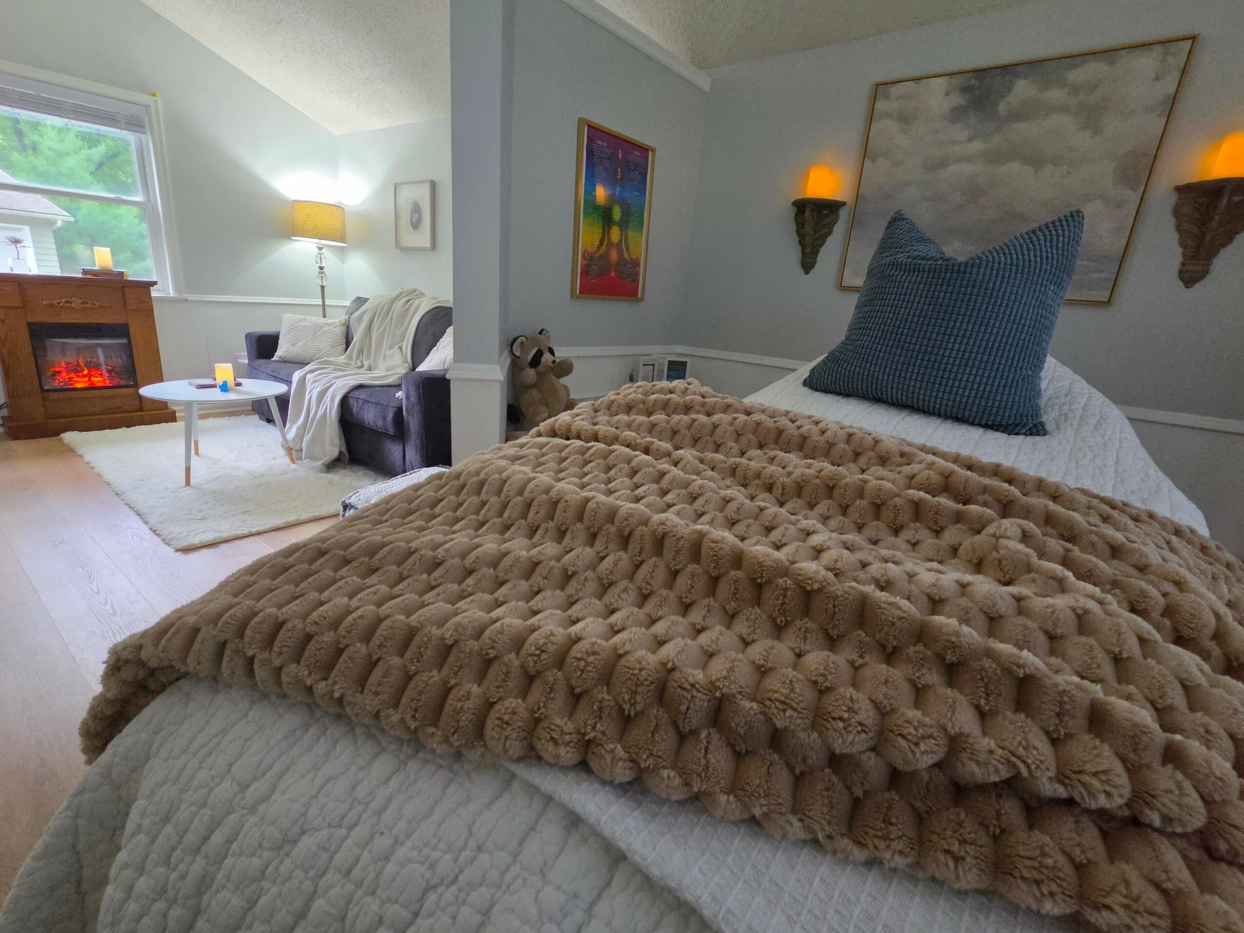 Bedroom with bed, blue pillow, wall art, and sitting area with fireplace, lamp, and window, in a cozy setting.