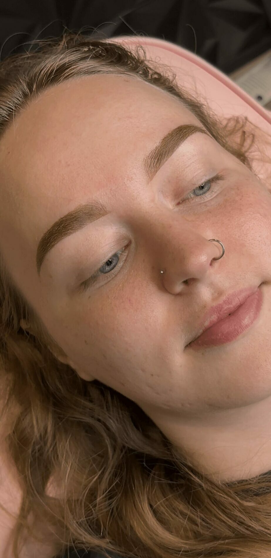 Close-up of a young woman's face with makeup, lying down with eyes closed, showing eyebrows, nose piercing, and relaxed expression.
