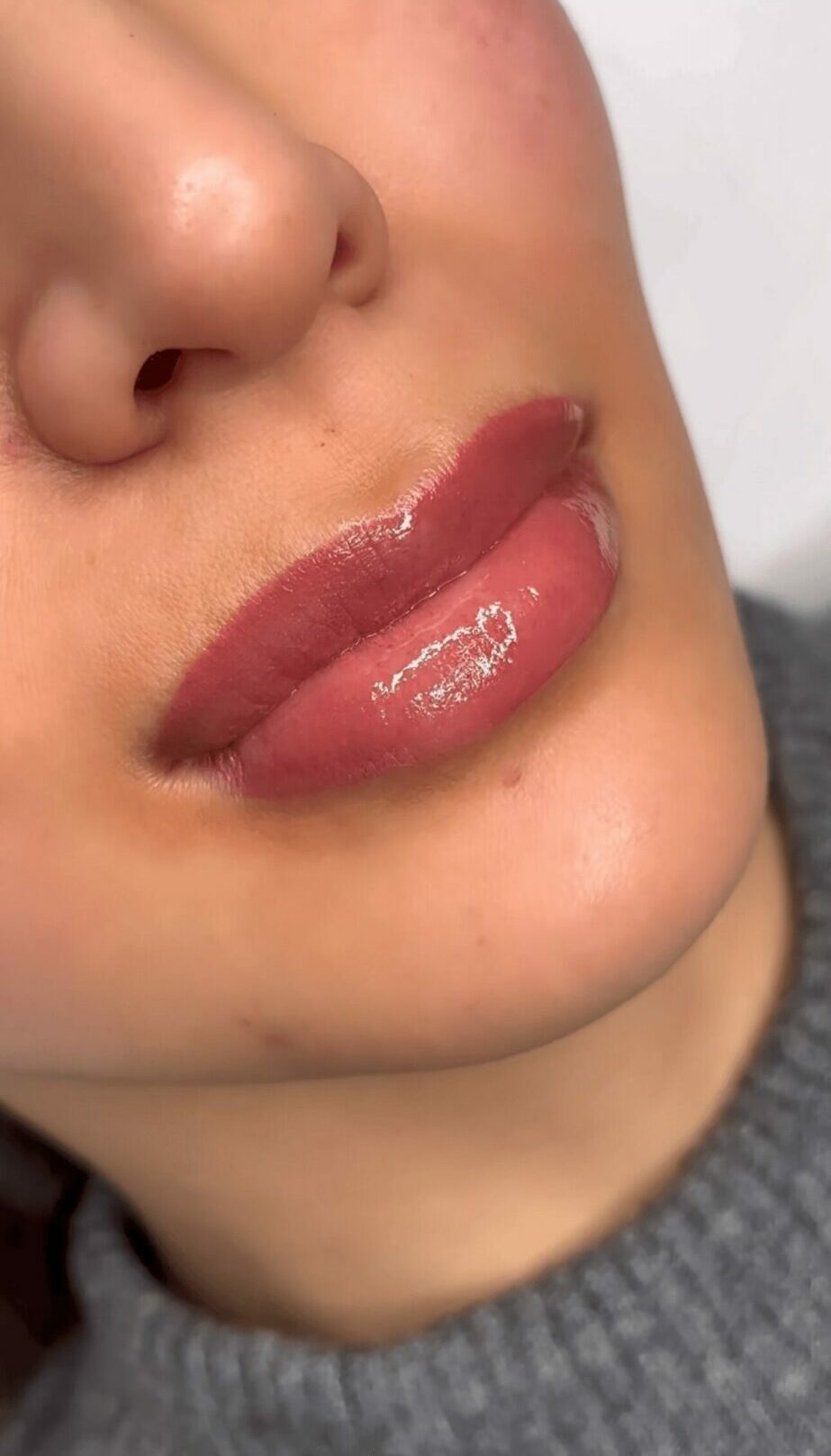 Close-up of a person's lips with glossy lipstick, showing skin and part of a gray sweater.