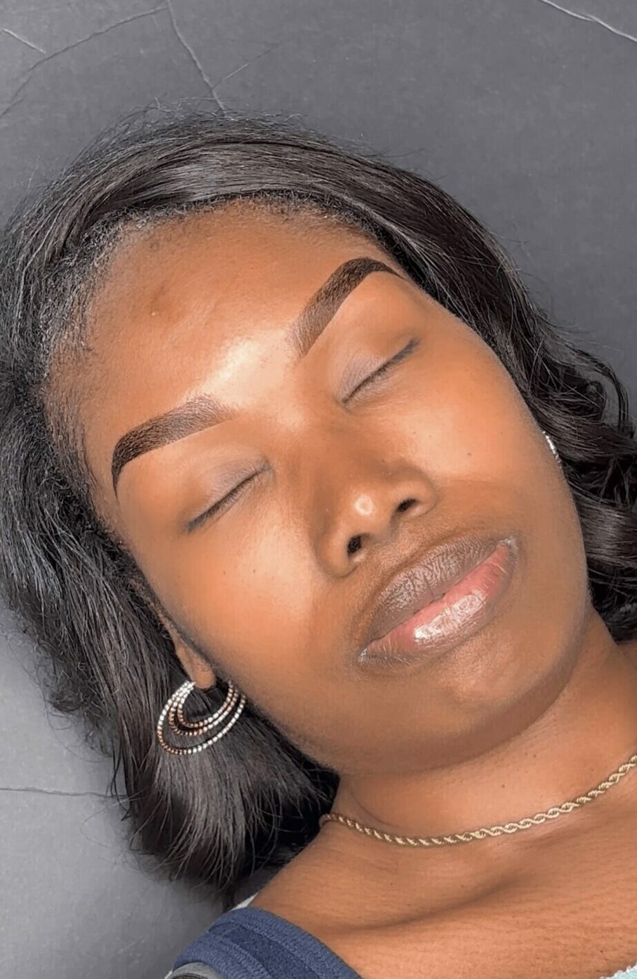 Close-up of a woman with closed eyes, wearing hoop earrings and a necklace, lying down with a neutral background.