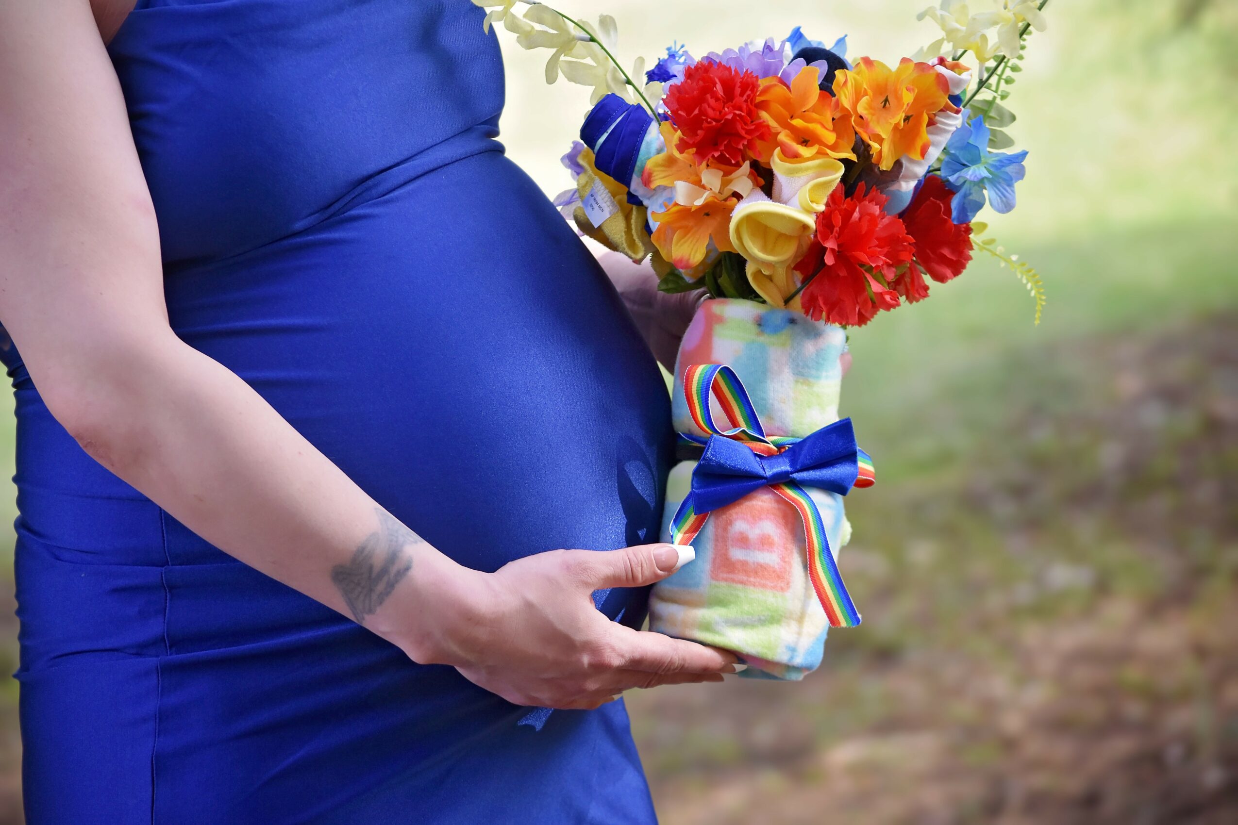Pregnant person in a blue dress holding a colorful bouquet of flowers in a vase with a ribbon, outdoors.