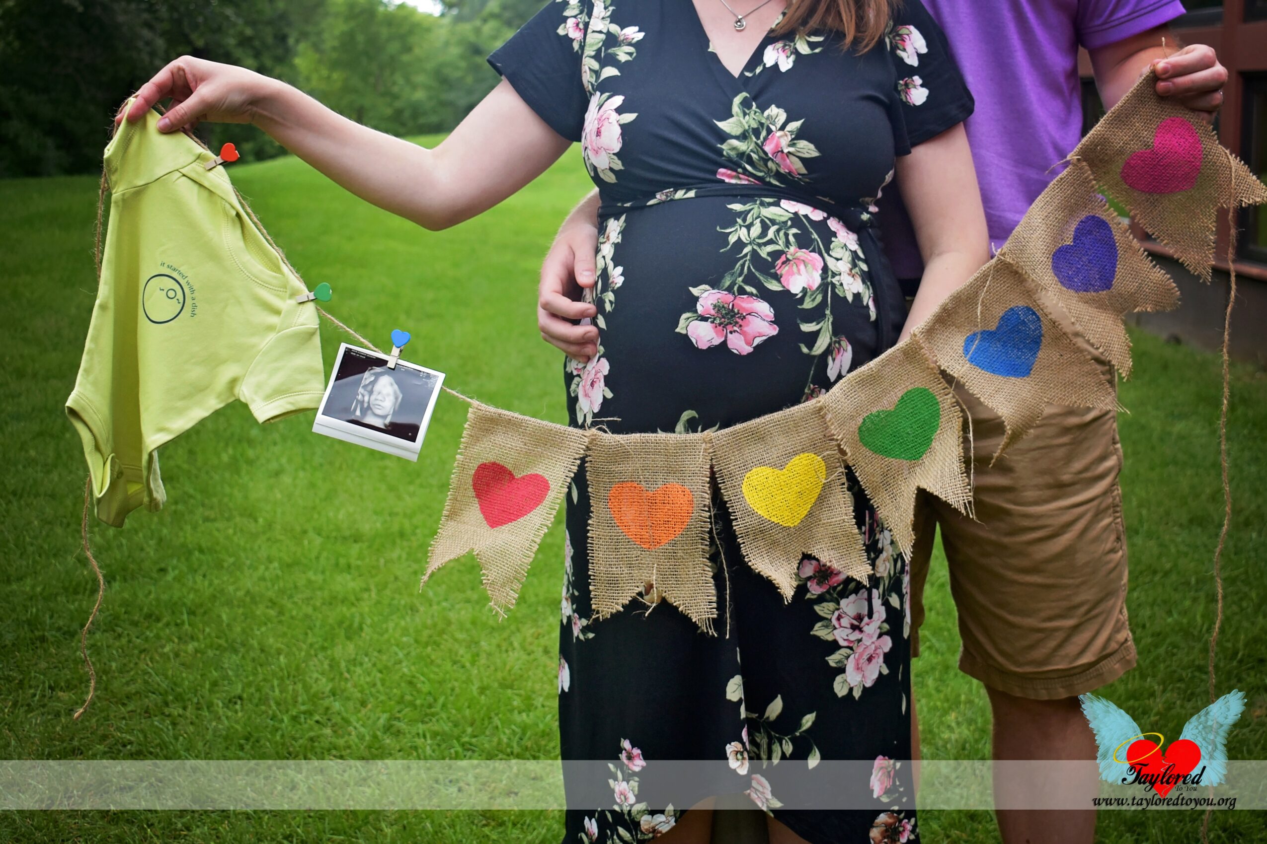 Pregnant woman holding a decorated banner with hearts outdoors, with a man behind her, green grass in background.