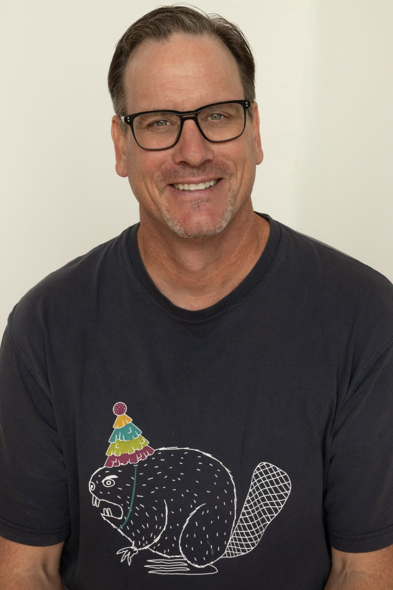 Man with glasses smiling, wearing a black T-shirt with a colorful animal graphic, against a plain background.