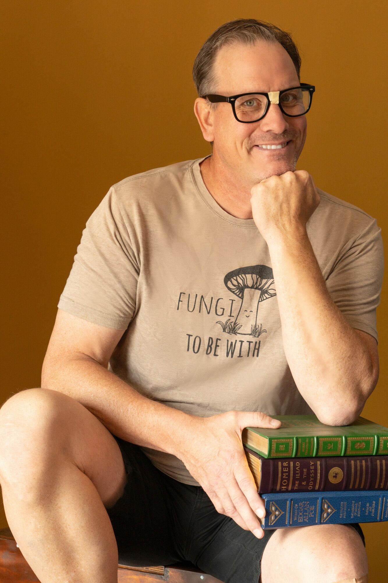 Man with glasses smiling, resting chin on hand, holding books, against a brown background.