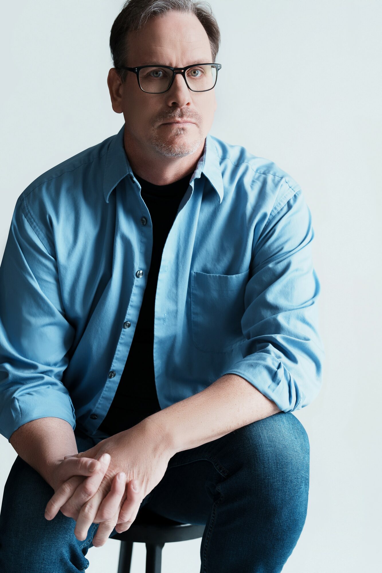 Man with glasses in a blue shirt sitting against a plain background, looking serious, with arms crossed.