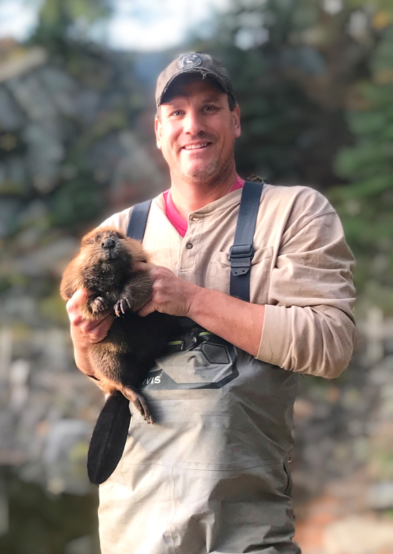 Man holding a small puppy outdoors with trees and rocks in background.