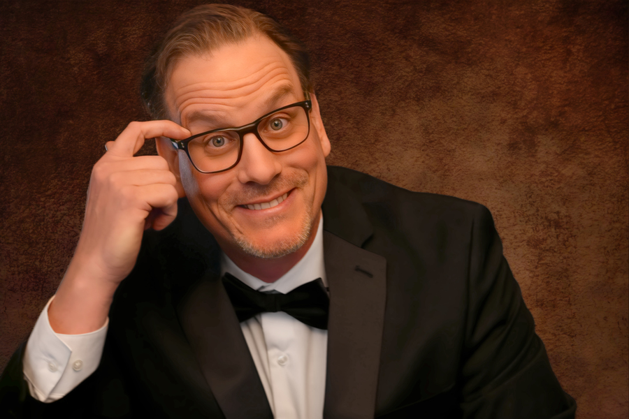 Man with glasses, smiling, adjusting his glasses, wearing a tuxedo, against a brown background.