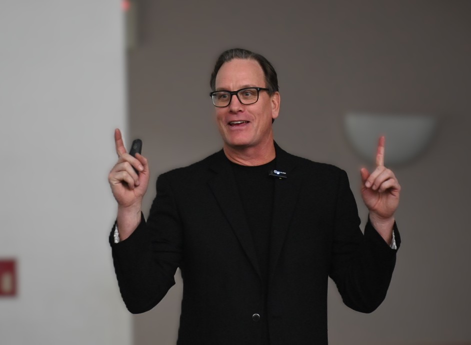 Man wearing glasses and black jacket speaking and gesturing with both hands indoors.
