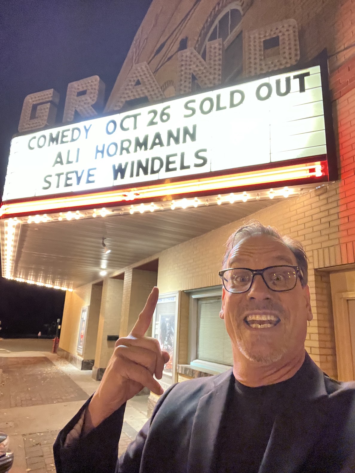 Man with glasses smiling and pointing upward outside theater with marquee sign, brick building in background.