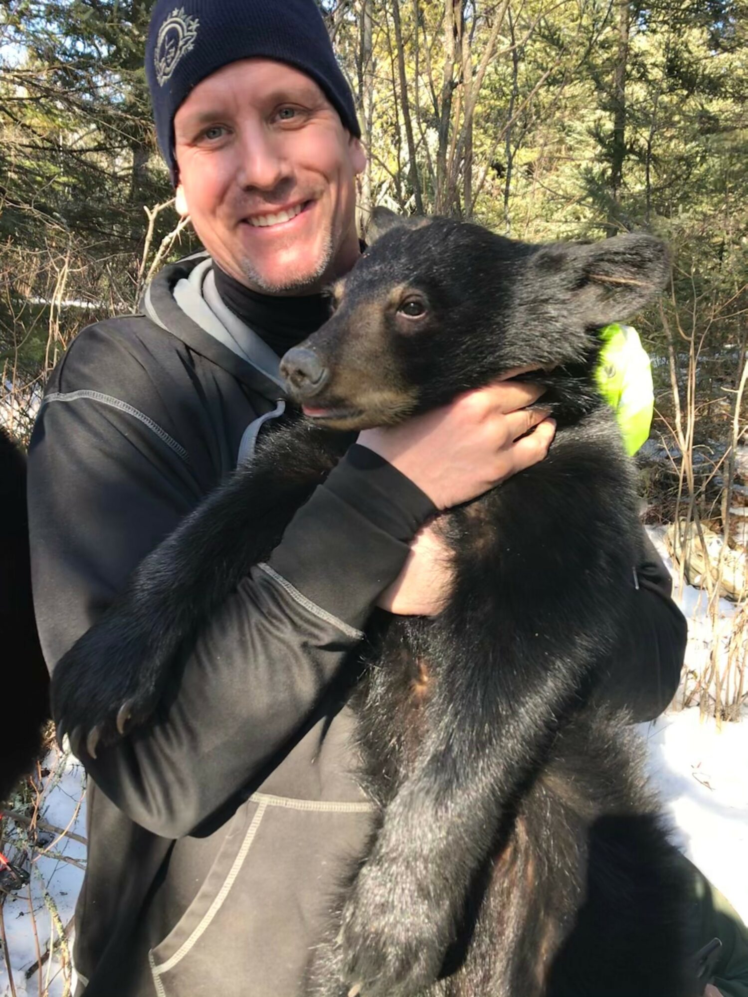 Man holding a black puppy outdoors in a wooded area, smiling, with sunlight filtering through trees.