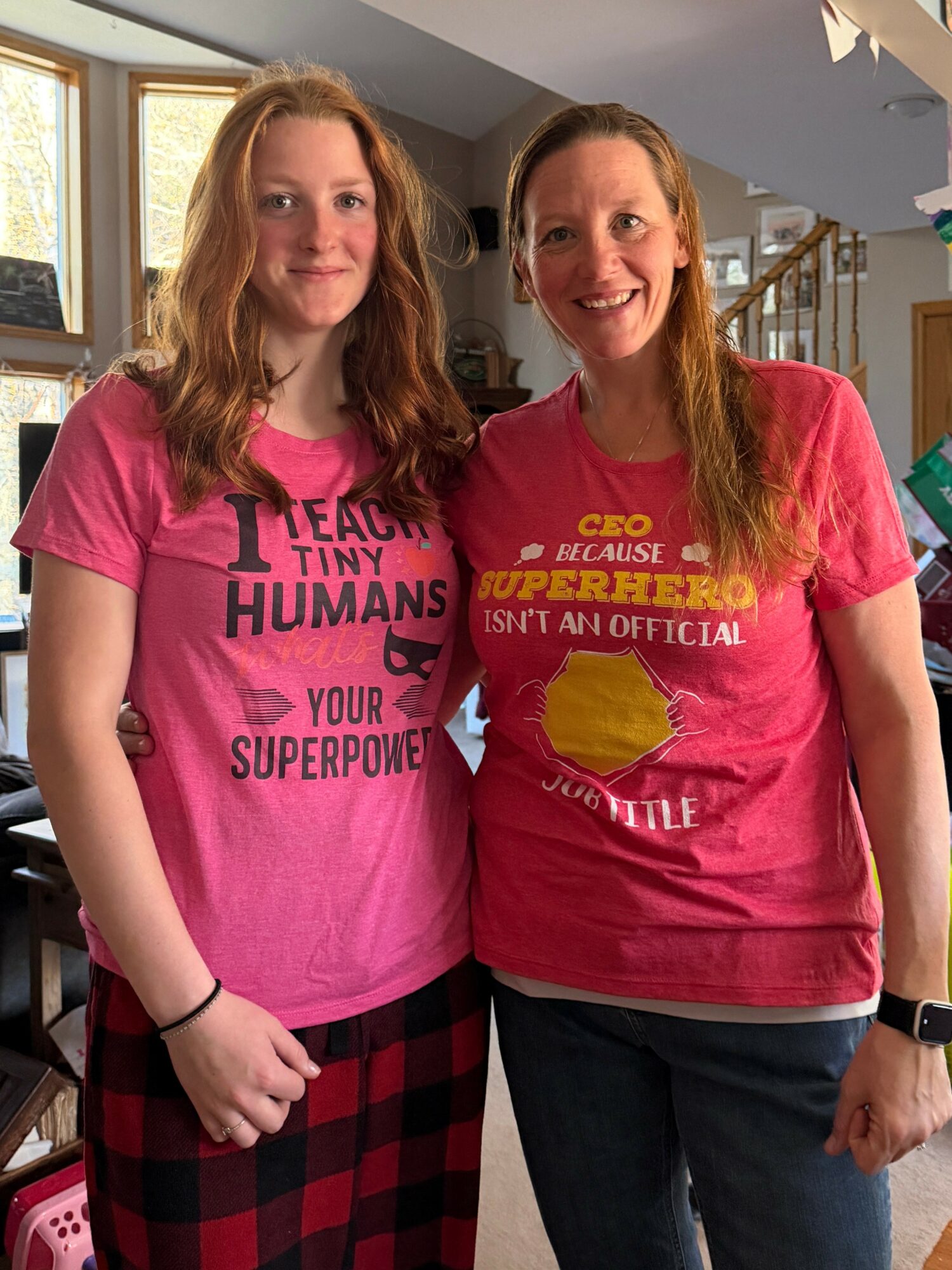 Two women standing indoors, smiling, wearing pink T-shirts with text, one with long red hair, the other with long light brown hair.