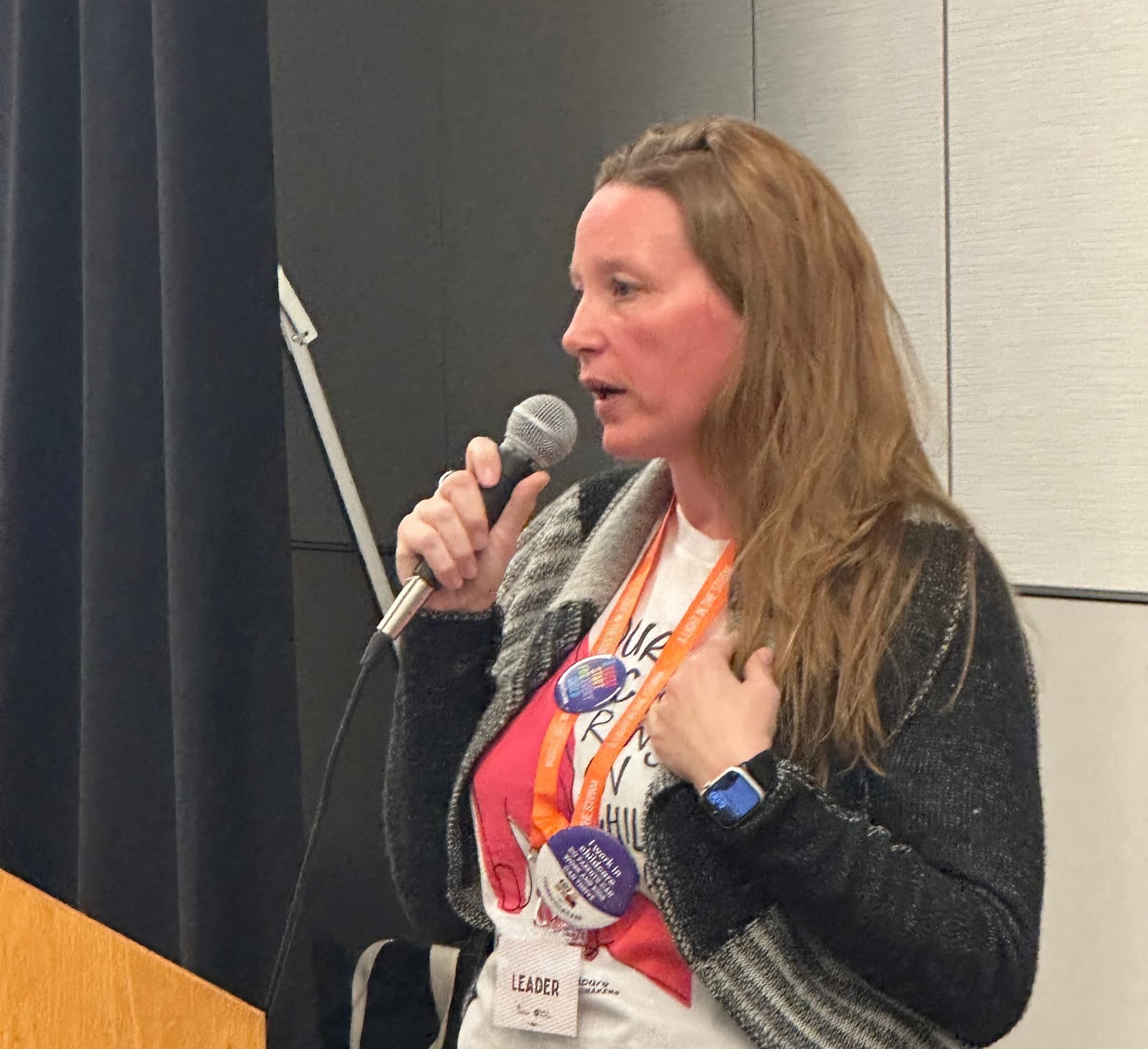 Woman speaking into a microphone at a conference, wearing a lanyard and a watch, with a dark background.