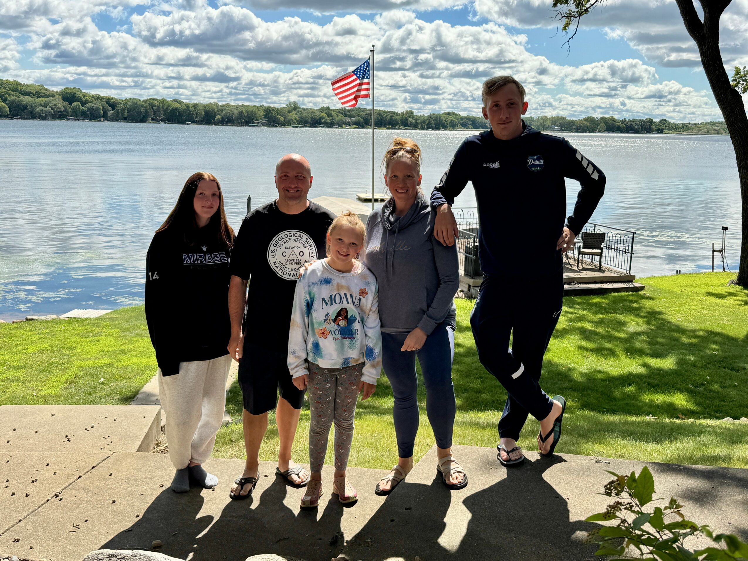 Group of five people standing by a lake with an American flag, trees, and a bench in the background.