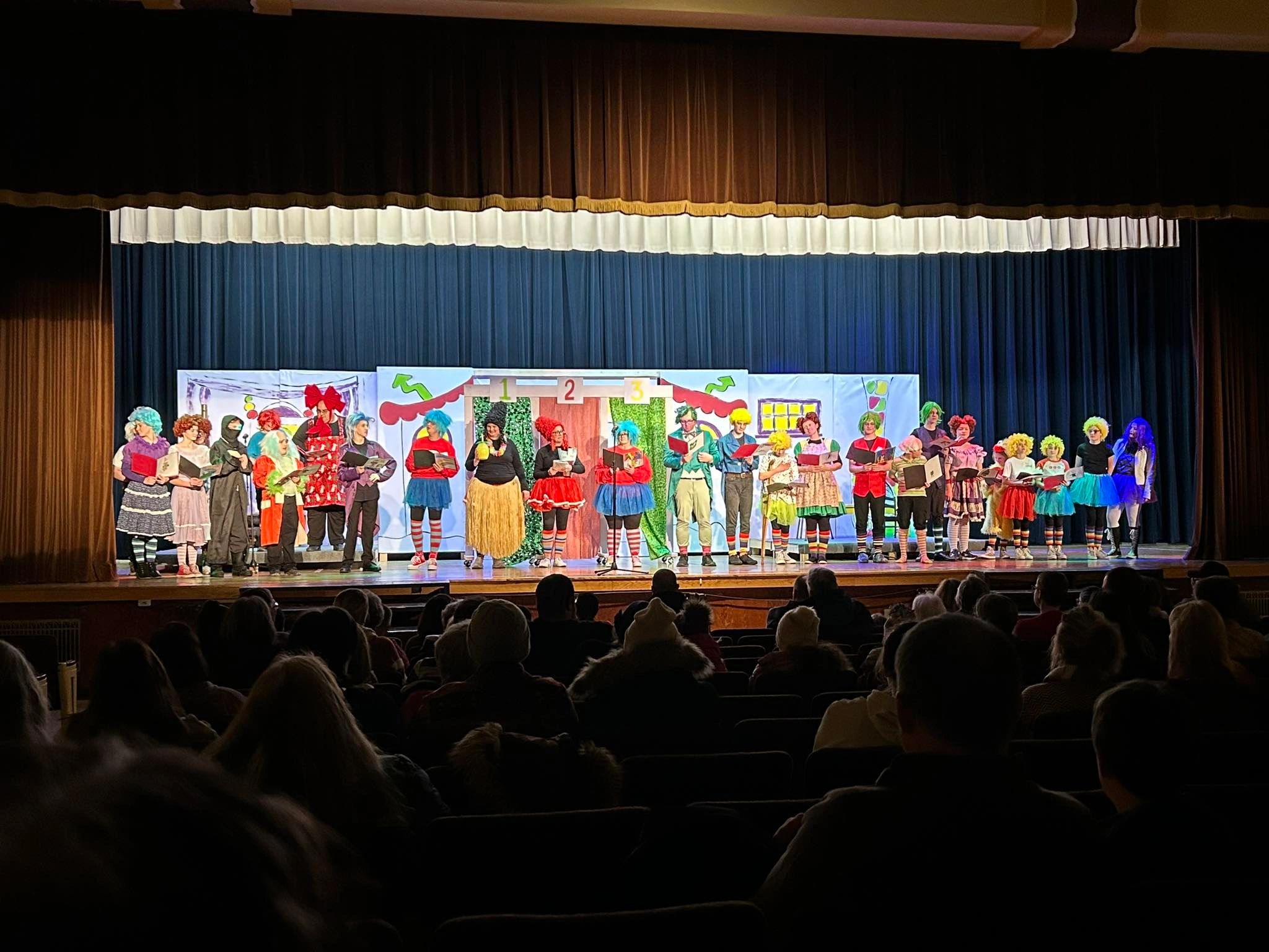 Children perform on stage with colorful backdrop and audience watching, stage curtains open, stage brightly lit.