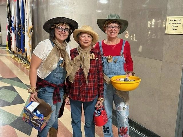 Three women dressed in cowboy-themed costumes, wearing hats and overalls, standing indoors with flags in the background.