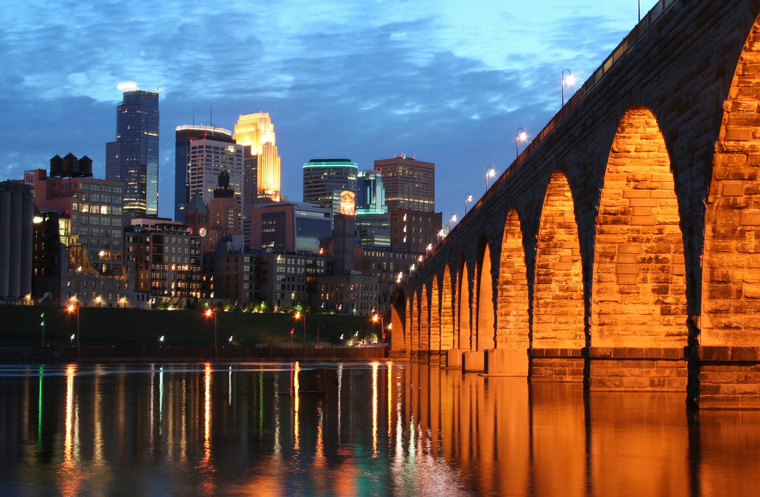 City skyline with illuminated buildings and a stone bridge reflecting in the water at dusk.